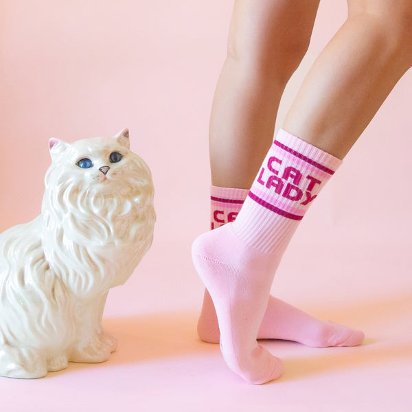 light pink cotton crew socks with ribbed cuffs and 'CAT LADY' text in magenta between pink stripes. Shown worn from the side next to white fluffy cat statue