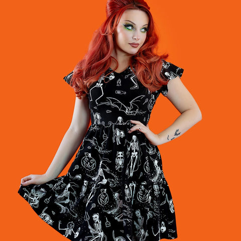 Model wearing a black cotton jersey knit dress with white skeleton, owl, pumpkin, witch and bat print. Dress has a shallow v-neckline, short flutter sleeves, a knee length double tiered skirt with ruffle hem and pockets. Shown from front against an orange background
