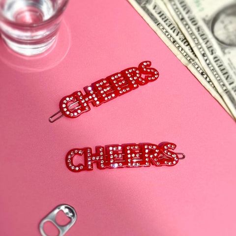 Red 'CHEERS' hair clips with clear rhinestone embellishments on a pink surface with money and a glass in the background