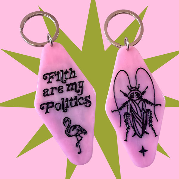 Two pink hotel-style keychains with silver metal key rings, displayed against a pink and green starburst background. One keychain features the black text 'Filth are my Politics' and a flamingo illustration, while the other features a black cockroach illustration.