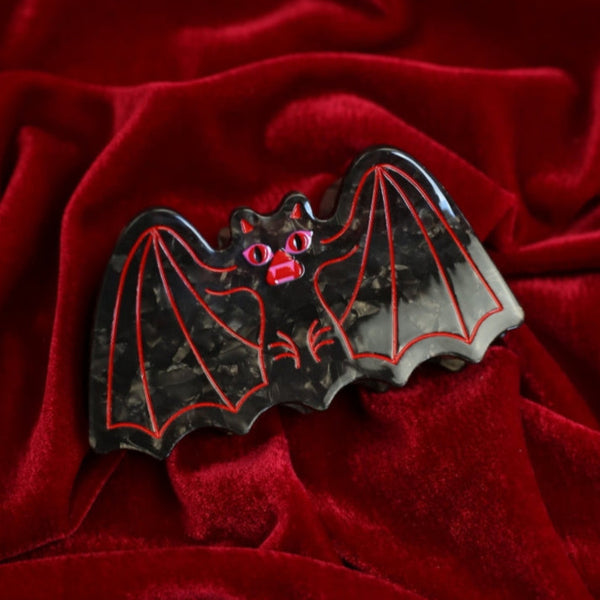 black marbled acetate claw style hair clip in the shape of a bat with outstretched wings and red and pink mouth with red line detail. Shown flat on red velvet background