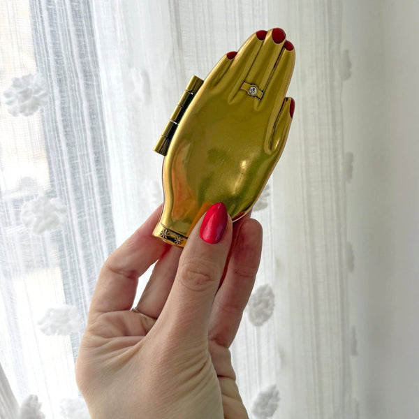bright slightly burnished finish gold hand-shaped compact mirror with red enameled manicure and clear jeweled ring and bracelet detail. shown closed held by hand for scale in front of white background