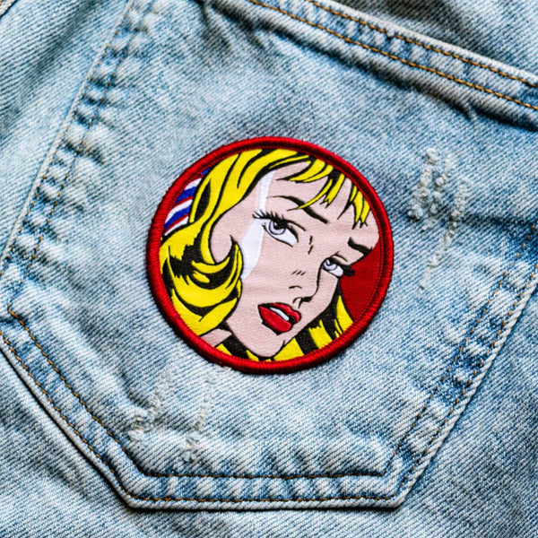 Circular patch with woven image of detail from Roy Lichtenstein painting of blonde woman on red background with red embroidered border. Shown on denim jeans