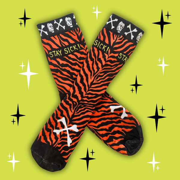 A pair of crew socks with a printed bold orange and black tiger stripe pattern, featuring white skulls and crossbones at the top, the text 'STAY SICK!' in yellow, and a white cross on the toe. The socks are arranged in an X shape on a bright lime green background decorated with black and white starburst graphics. 