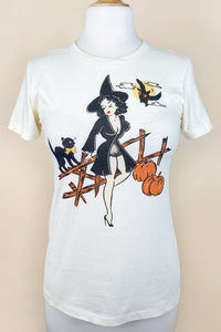 off-white womens cut t-shirt with illustrated graphic of pinup style woman wearing black negligee and witch hat resting on a wooden fence with hissing black cat, owl, and two pumpkins. Shown untucked on a dress form