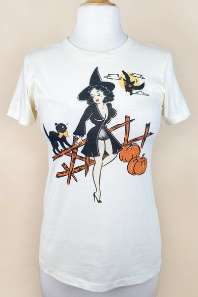 off-white womens cut t-shirt with illustrated graphic of pinup style woman wearing black negligee and witch hat resting on a wooden fence with hissing black cat, owl, and two pumpkins. Shown untucked on a dress form