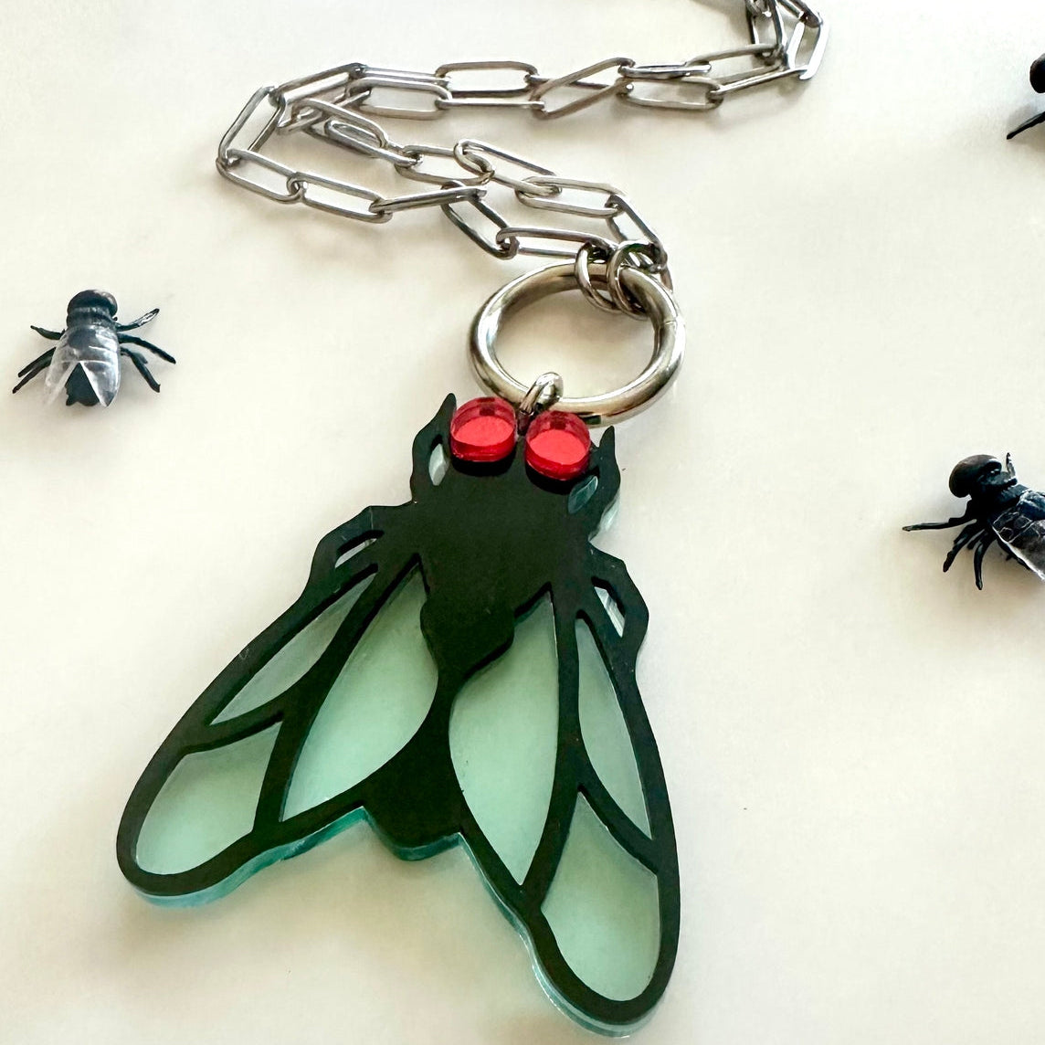 Laser-cut acrylic in the shape of a house fly with black body and transparent matte finish blue wings and red mirrored eyes. Attached to silver metal paperclip style chain via silver metal O-ring. Shown on white background with plastic toy flies