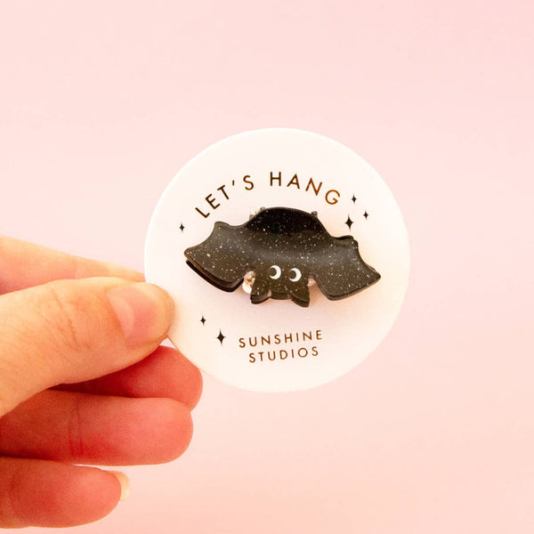 A black and silver glitter acrylic hair clip in the shape of a bat with big translucent white eyes. Shown on packaging card