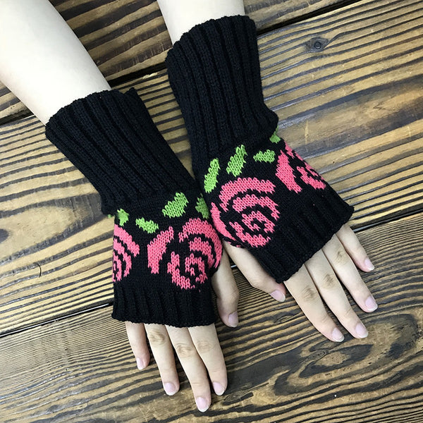 knit black fingerless gloves with ribbed cuffs and a knit-in pattern of pink roses with green leaves. Shown on a model with palms facing downward on a wood background