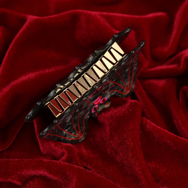 black marbled acetate claw style hair clip in the shape of a bat with outstretched wings and red and pink mouth with red line detail. Shown from bottom on red velvet background to display metal hardware