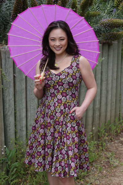 Model wearing a sleeveless stretch cotton dress in a black background with pink, purple, and creamy off-white flowers with goldenrod colored stems. Dress has sweetheart neckline, fitted bodice with vertical seaming, full gathered knee length skirt and pockets