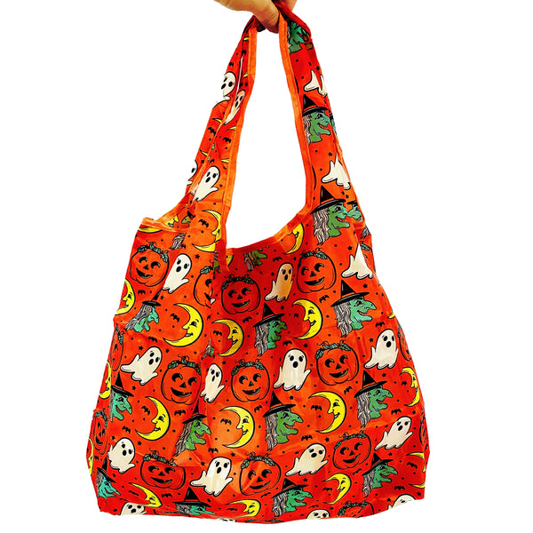 An orange reusable nylon tote bag with an all-over pattern of cartoonish retro Halloween witches, ghosts, crescent moons, and jack-o'-lanterns shown held by hand on white background