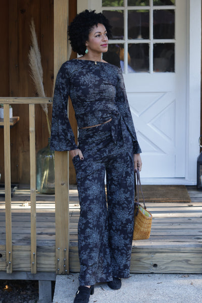 Model wearing stretch cotton knit high-waisted wide leg pants in a dark blue background with dainty floral pattern of heather shrub and its leaves. Pants have a wide waistband and pockets. Shown standing in front of cabin door
