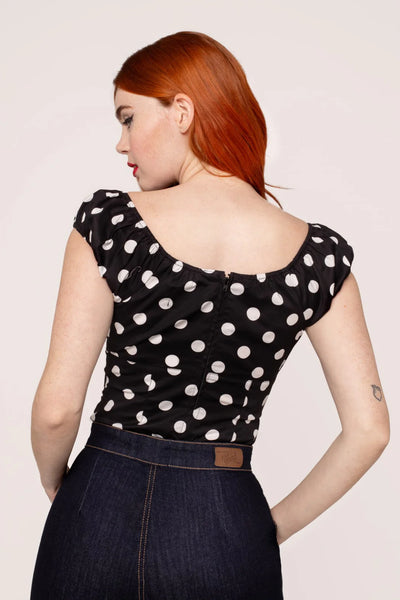 model wearing woven short sleeve top in black with large white polka dot pattern. Top has round elasticized neckline and cuffs, keyhole detail with red heart-shaped button, and underbust seam. Shown from back tucked into jeans