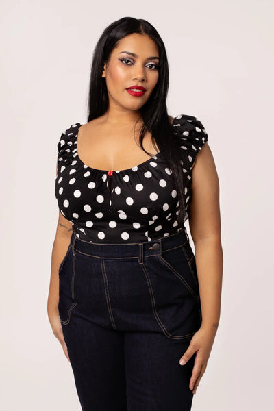 plus size model wearing woven short sleeve top in black with large white polka dot pattern. Top has round elasticized neckline and cuffs, keyhole detail with red heart-shaped button, and underbust seam. Shown from front tucked into jeans
