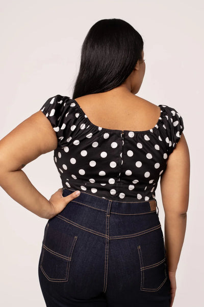 plus size model wearing woven short sleeve top in black with large white polka dot pattern. Top has round elasticized neckline and cuffs, keyhole detail with red heart-shaped button, and underbust seam. Shown from back tucked into jeans