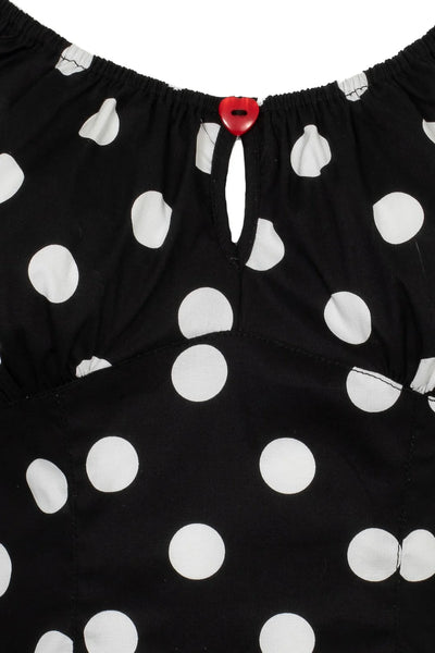 woven short sleeve top in black with large white polka dot pattern. Top has round elasticized neckline and cuffs, keyhole detail with red heart-shaped button, and underbust seam. Shown from front in detail close up of bodice