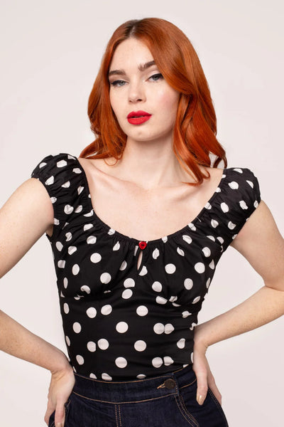 model wearing woven short sleeve top in black with large white polka dot pattern. Top has round elasticized neckline and cuffs, keyhole detail with red heart-shaped button, and underbust seam. Shown from front tucked into jeans