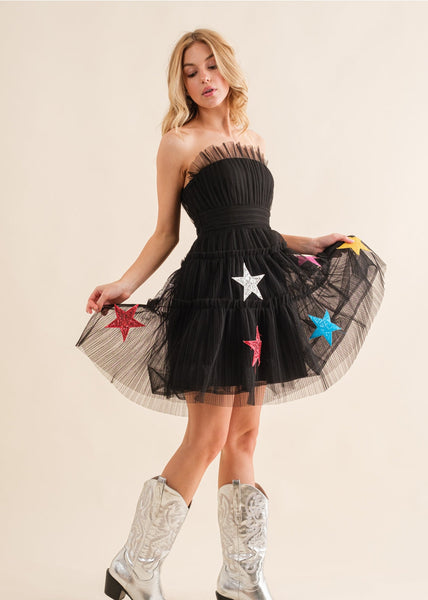black mini dress with a straight neckline made from layers of tulle with multicolored sequined star appliques on the bottom two tiers of the dress. The dress has a tulle banded waist. Shown on a model from side