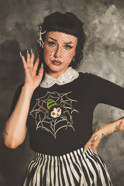 black crew neck short sleeved sweater with white, bright orange, green, and black printed image of cat with spider body and legs smiling on top of a spider web. Shown on model styled with white crochet collar