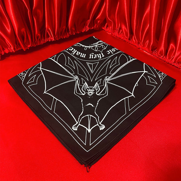 Square black cotton bandana with screen printed white image of mirrored bats and spiders inside a web with center image of vampire skull with quote “ Listen to them, the children of the night. What music they make!”. Shown folded to show only bat printed corner on red velvet background