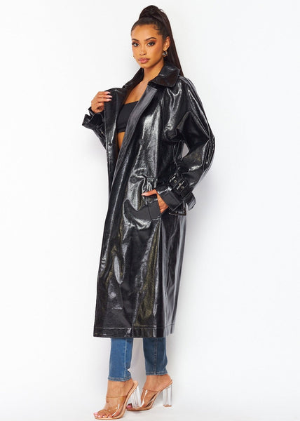 model wearing oversized shiny textured pvc black trench coat with notched collar, relaxed shoulders, self belt with self buckle and oversized buckles at cuffs, double breasted button closure, and slash pockets. Shown from side with coat open