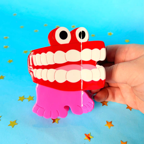 large cellulose acetate claw-style hair clip in the shape of wind-up chattering teeth with a red mouth, white teeth, black eyes, and pink feet, held by a hand against a bright blue background with scattered gold star confetti.