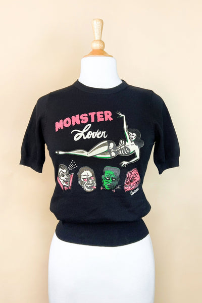 black crew neck short sleeved sweater with 'Monster Lover' text in pink and white and illustration of pinup style woman in black and white skeleton print swim suit and oversized heads of a vampire, wolfman, Frankenstein, and gorilla with hearts in their eyes. Shown on dress form