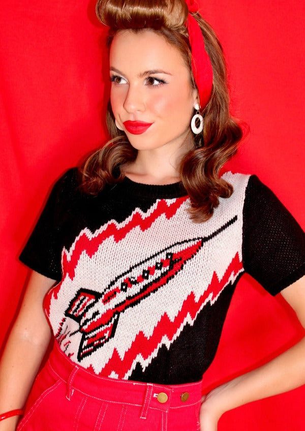 Model wearing a fitted acrylic short sleeve sweater with a retro looking rocket ship on a red, white, and black background.