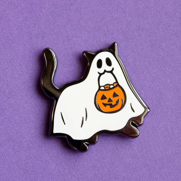 Silver metal enamel pin of a black cat wearing a ghost sheet costume with jack-o’-lantern shaped basket in its mouth. Shown on purple background 