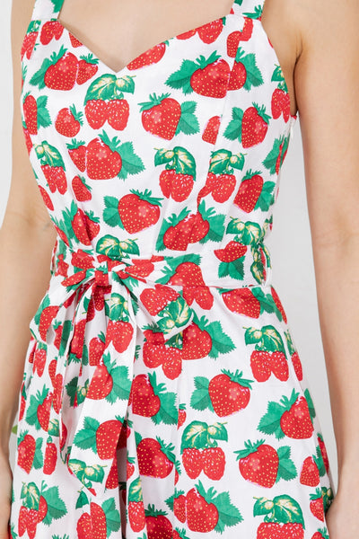 Model wearing sleeveless fit and flare cotton dress with white background all-over pattern of strawberries and raspberries with green leaves. It has a sweetheart neckline, wide straps, princess seaming at the bodice, a self tie at the waist and a flared knee length skirt with pockets. Shown from the front In close up of bodice and waist for pattern detail