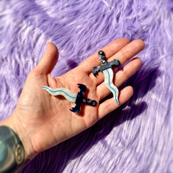 pair of acetate hair clips in the shape of small daggers with wavy iridescent blades and marbled blue and white handles with a row of three rhinestones in each. Shown held in an open palm on a purple faux fur background