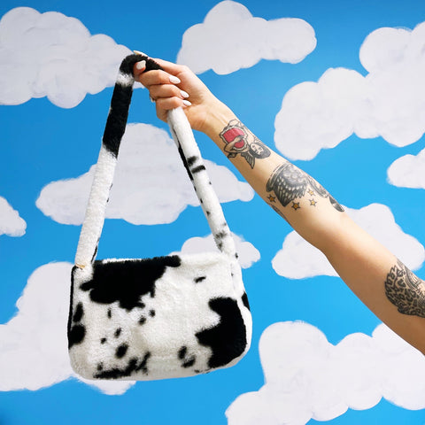 Plush black and white cow print faux fur purse with shoulder strap seen held by a model in front of a blue sky and white cloud background 