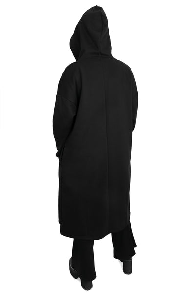 model wearing a black fleece longline open front hoodie sweater with oversized hood with drawstring, long sleeves with thumbholes, and pockets. Shown from back with hood up and hands in pockets