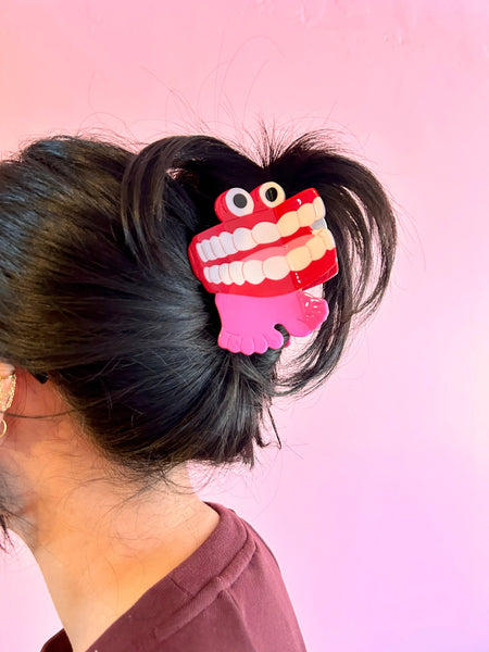 large cellulose acetate claw-style hair clip in the shape of wind-up chattering teeth with a red mouth, white teeth, black eyes, and pink feet, shown worn in an updo from the side
