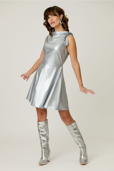 Model wearing a metallic silver knit fit and flare mini dress with cowl collar neck with asymmetrical self tie detail, darting at bust, and above the knee length skirt with pockets. Shown from front