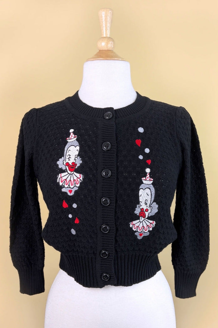 black cotton pointelle knit cardigan with embroidery detail on both sides of the chest of two white, red, and grey clowns surrounded by red hearts and white circles. Shown on a dress form