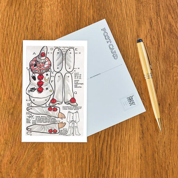 postcard with image of instructions for assembling a clown doll with detailed illustrations and text on wooden surface with pen