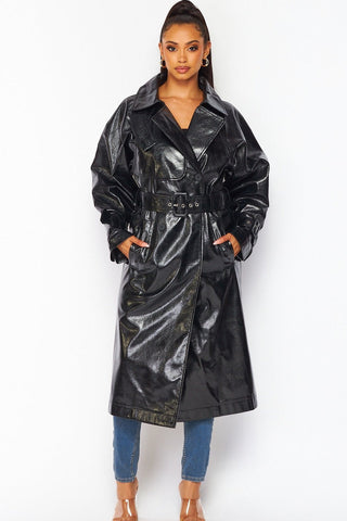 model wearing oversized shiny textured pvc black trench coat with notched collar, relaxed shoulders, self belt with self buckle and oversized buckles at cuffs, double breasted button closure, and slash pockets. Shown from front with coat buckled