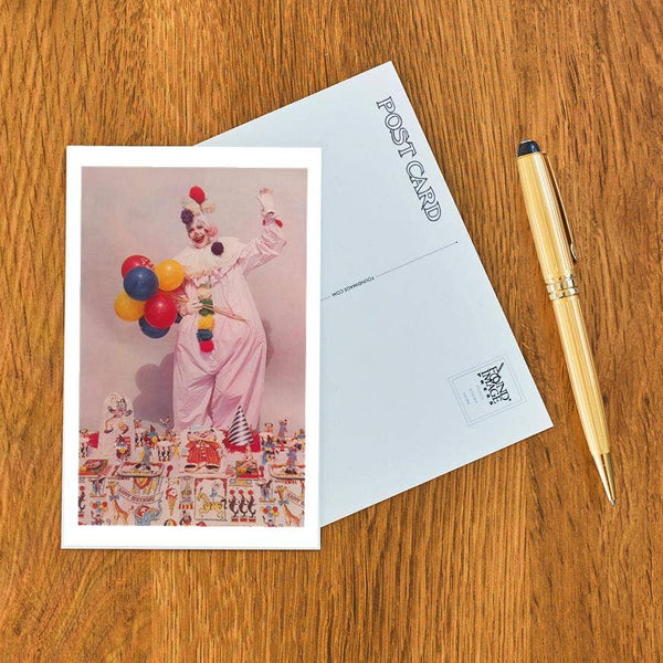 postcard of a vintage color photograph of a clown holding balloons in front of a collection of circus themed toys on a wooden surface with a pen.