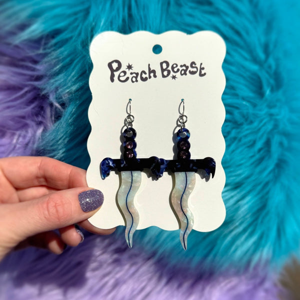 pair of acetate dangle earrings in the shape of small daggers with wavy iridescent blades and marbled blue and white handles with a row of three rhinestones in each. Shown on packaging card