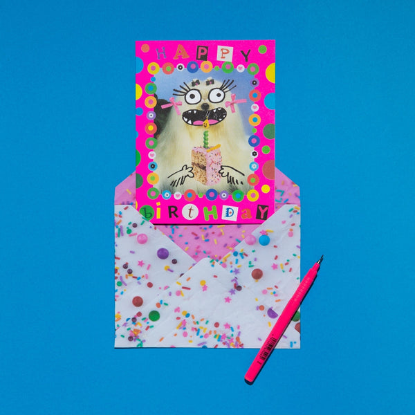 "Happy Birthday" message collage card with neon pink background & polka dots with photo of Maltese dog with added cartoon face blowing out the candle on a slice of birthday cake. Shown with frosting and sprinkles patterned envelope on blue background