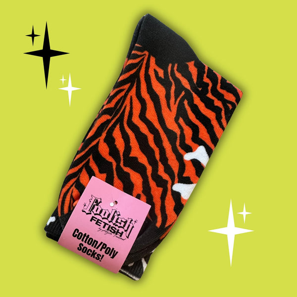 A pair of crew socks with a printed bold orange and black tiger stripe pattern, featuring white skulls and crossbones at the top, the text 'STAY SICK!' in yellow, and a white cross on the toe. The socks are shown in their branded pink packaging card