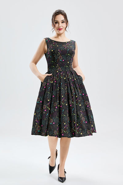 Model wearing a fit and flare dress made of soy silk in a black background pattern of exploding fireworks in bright red, pink, green, blue, orange, and purple. It has a high fitted neckline, princess seamed bodice, wide banded waist, and box pleated knee length skirt with pockets. Shown from the front