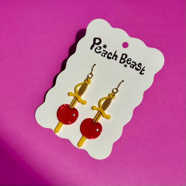 acetate dangle earrings in the shape of a pair of yellow cocktail sword skewers with a marbled maraschino cherry with silver rhinestone decoration. Shown flat in packaging card