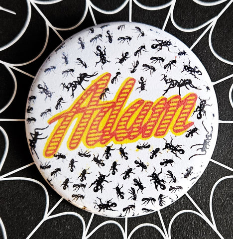 1.25” round pinback button with logo for Adam Ant in yellow and red striped script surrounded by small black ants on white background 