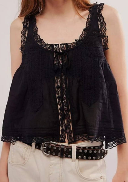 model wearing black cotton sleeveless camisole top with round neckline, floral lace ruffle at straps and bottom hem, tie detail at center with small plastic buttons running down bodice, vertical pin tuck detail at top of bodice and horizontal pin tuck detail at bottom hem, and geometric lace running vertically down each side of top