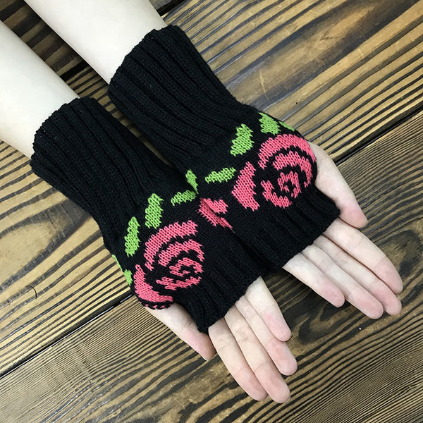 knit black fingerless gloves with ribbed cuffs and a knit-in pattern of pink roses with green leaves. Shown on a model with palms facing upward on a wood background