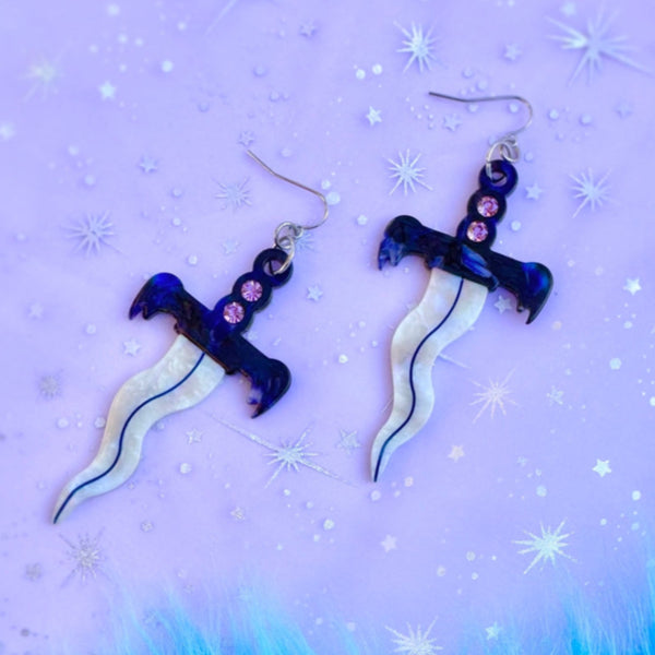 pair of acetate dangle earrings in the shape of small daggers with wavy iridescent blades and marbled blue and white handles with a row of three rhinestones in each. Shown flat on purple sparkle backgrounf
