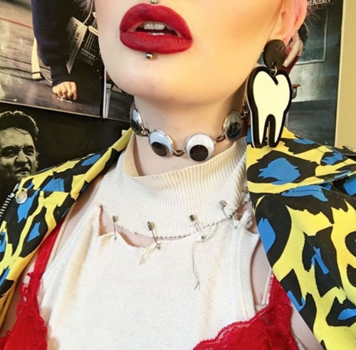 Model wearing choker style necklace with 8 linked large plastic black and white googly eyes with gunmetal finish metal link chain extender and matching swivel hook clasp closure. Shown worn with leopard print dress shirt and tooth shaped earrings